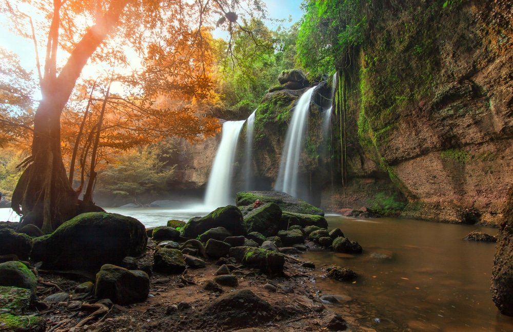 น้ำตกไหลผ่านหน้าผาที่ปกคลุมด้วยมอส ล้อมรอบด้วยหิน ต้นไม้ และความเขียวขจี ณ สถานที่ท่องเที่ยวเขาใหญ่ โดยมีแสงแดดส่องผ่านใบไม้ลงมา.