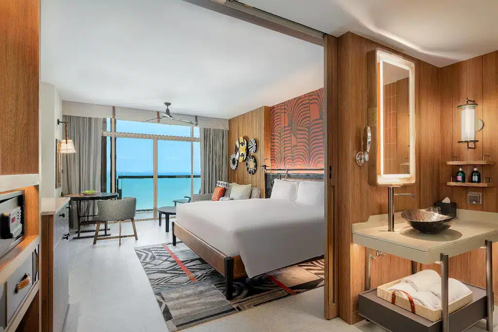 ห้องพักโรงแรมสมัยใหม่พร้อมเตียงขนาดใหญ่, มุมนั่งเล่น, โต๊ะทำงาน, และห้องน้ำแบบเปิดโล่ง; หน้าต่างบานใหญ่จากพื้นจรดเพดานให้ทัศนียภาพของทะเล—เหมาะสำหรับผู้ที่ต้องการ พักพัทยาติดทะเล อย่างสบายและมีสไตล์.