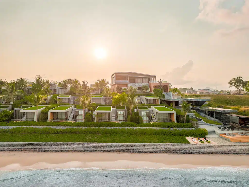 รีสอร์ทริมชายหาดทันสมัยพร้อมวิลล่าหลายหลัง หลังคาสีเขียว ต้นปาล์ม และสวนที่จัดแต่งอย่างสวยงาม เหมาะสำหรับการพักผ่อนริมทะเลพัทยา มองเห็นวิวพระอาทิตย์ตกจากชายฝั่ง.