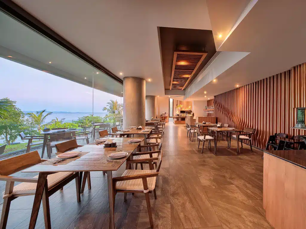ร้านอาหารสมัยใหม่พร้อมโต๊ะและเก้าอี้ไม้ หน้าต่างบานใหญ่จากพื้นจรดเพดานที่มอบวิวทิวทัศน์ของต้นไม้และน้ำ พร้อมการออกแบบภายในที่ทันสมัยและแสงไฟอบอุ่น—เหมาะสำหรับผู้ที่ต้องการ พักพัทยาติดทะเล อย่างมีสไตล์.