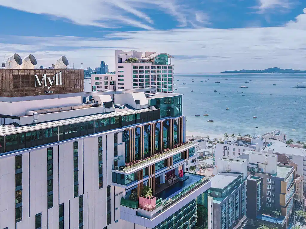โรงแรมสูงสมัยใหม่ที่ชื่อว่า "Mytt Hotel" ตั้งอยู่เหนือชายหาดที่มีเรือลำน้อยลอยอยู่ในน้ำ ให้บริการ พักพัทยาติดทะเล พร้อมวิวเมืองใกล้เคียงภายใต้ท้องฟ้าที่มีเมฆบางส่วน.