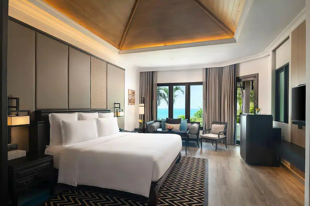 ห้องพักโรงแรมสมัยใหม่พร้อมเตียงขนาดใหญ่ พื้นที่นั่งเล่น โทรทัศน์จอแบน และพื้นไม้ ให้ทัศนียภาพอันงดงามของทะเลและต้นปาล์มผ่านหน้าต่างบานกว้าง—เหมาะสำหรับผู้ที่ต้องการ พักพัทยาติดทะเล อย่างสะดวกสบายและมีสไตล์.