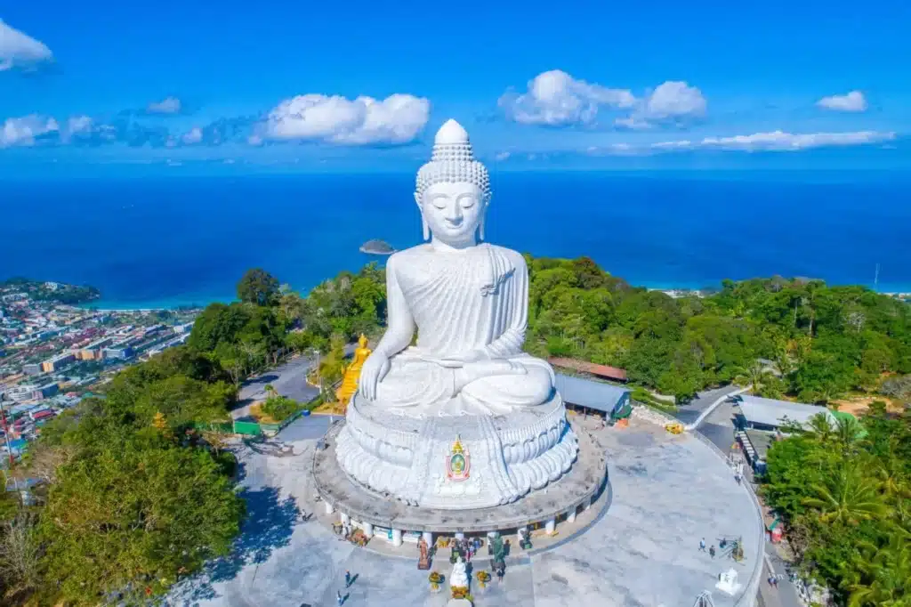 มุมมองทางอากาศของพระพุทธรูปสีขาวขนาดใหญ่ที่วัดรีภูเก็ตนั่งอยู่บนยอดเขาที่เขียวขจี เมืองชายฝั่ง และมหาสมุทรเป็นพื้นหลังภายใต้ท้องฟ้าสีฟ้าใส ที่เที่ยวภูเก็ต