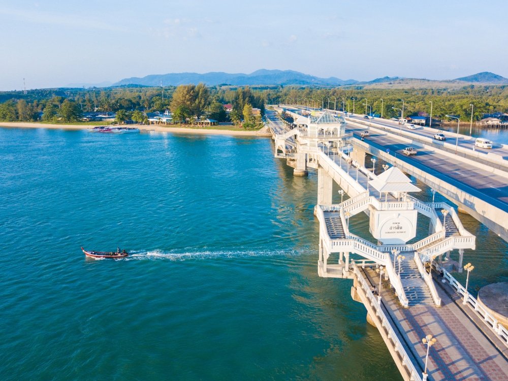 มุมมองทางอากาศของสะพานข้ามน้ำที่มีเรือเล็กแล่นลอดใต้ เชื่อมแนวชายฝั่งที่มีต้นไม้ เที่ยวภูเก็ต ทิวทัศน์ใกล้กับวัดเลภูเก็ตหลายแห่ง