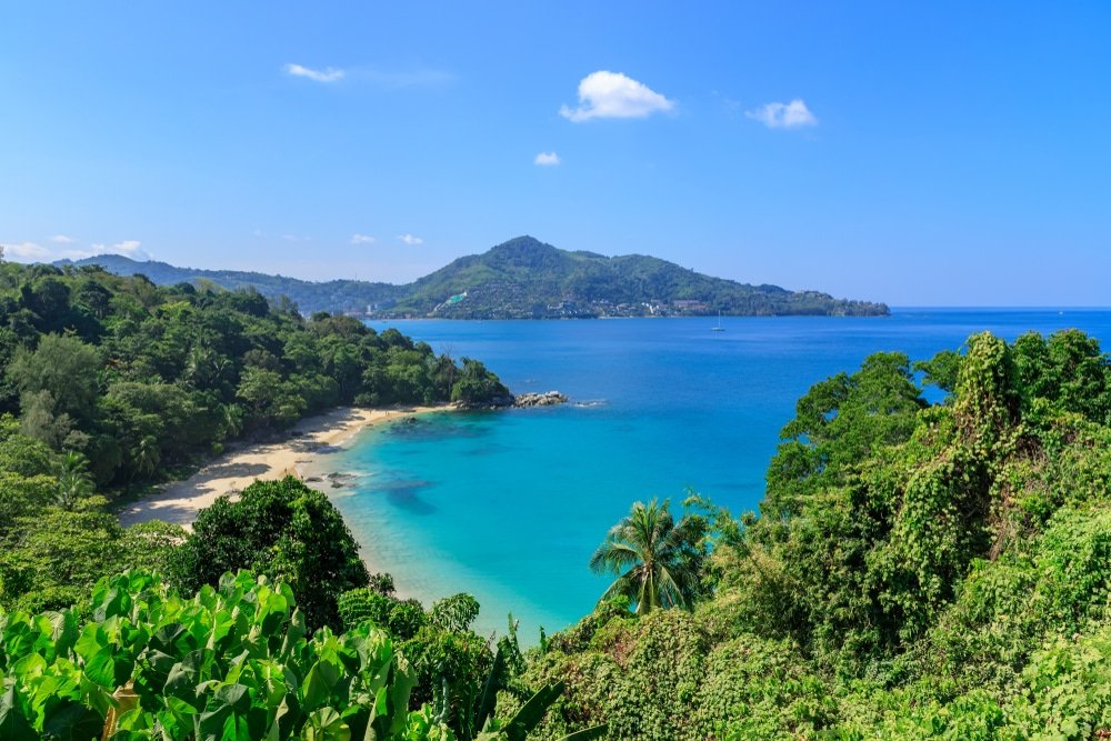 ทิวทัศน์อันงดงามของแนวชายฝั่งเขตร้อนที่มีหาดทราย พืชพรรณสีเขียวชอุ่ม  ที่เที่ยวภูเก็ต และน้ำทะเลสีฟ้าใสทอดยาวไปจนถึงเกาะที่อยู่ห่างไกลภายใต้ท้องฟ้าสีฟ้าสดใส เสริมด้วยความงามอันเงียบสงบของวัดภูเก็ตในบริเวณใกล้เคียง