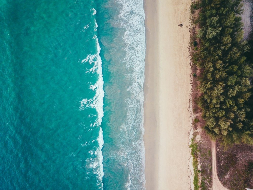 มุมมองทางอากาศของชายหาดในภูเก็ตแสดงให้เห็นคลื่นสีฟ้าคราม ท่องเที่ยวภูเก็ต หาดทรายกว้าง และความเขียวขจีหนาแน่นทางด้านขวา โดยมีวัดจังหวัดภูเก็ตตั้งอยู่ท่ามกลางต้นไม้