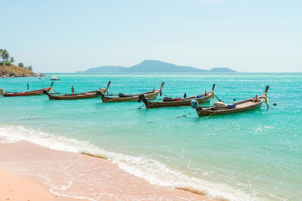 เรือหางยาวหลายลำจอดอยู่ในน้ำทะเลสีฟ้าครามใกล้หาดทราย ที่เที่ยวภูเก็ต โดยมีเกาะที่มองเห็นได้แต่ไกลภายใต้ท้องฟ้าที่แจ่มใส ทำให้มองเห็นทิวทัศน์ที่ชวนให้นึกถึงวัดเลภูเก็ต