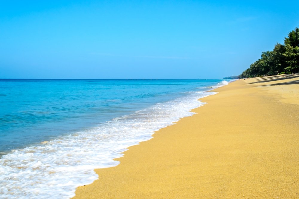 ฉากชายหาดอันเงียบสงบที่มีน้ำทะเลสีฟ้าใสและหาดทรายสีทอง ที่เที่ยวภูเก็ต ล้อมรอบด้วยแนวต้นไม้สีเขียวภายใต้ท้องฟ้าที่แจ่มใส ชวนให้นึกถึงความสงบสุขที่พบได้ใกล้กับวัดรีสอร์ตภูเก็ต