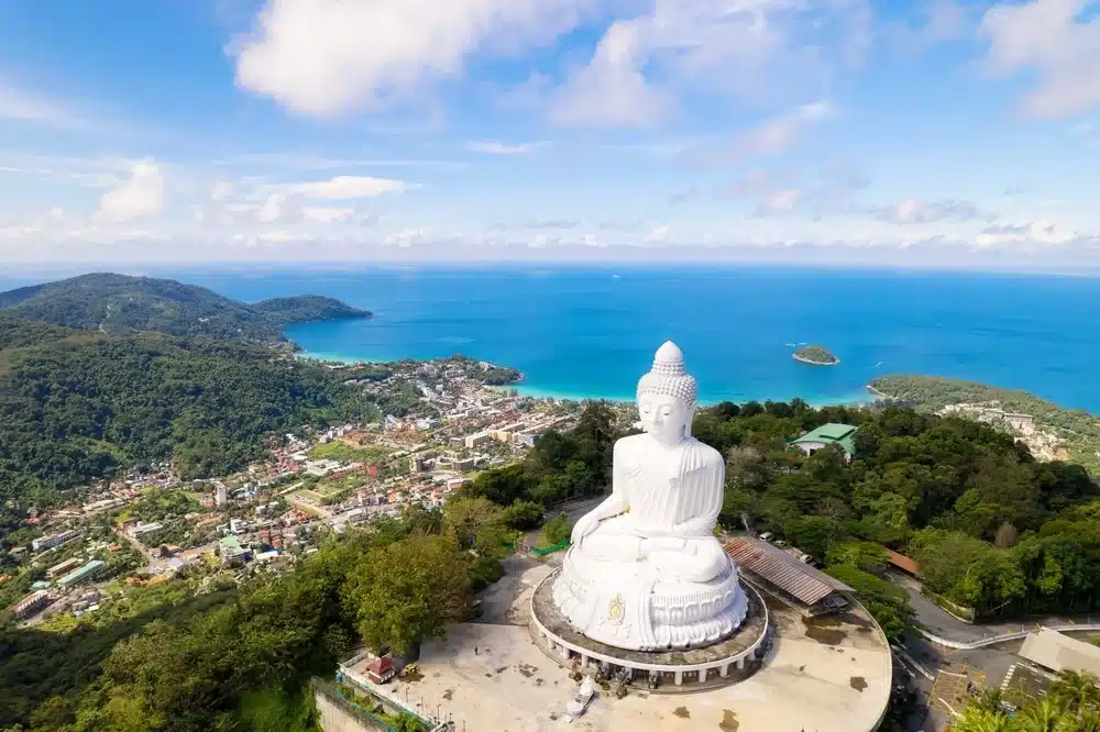 มุมมองทางอากาศของพระใหญ่บนยอดเขาที่มองเห็นความเขียวขจี เมืองด้านล่าง และทะเลสีฟ้าพร้อมท้องฟ้า 
 ที่เที่ยวภูเก็ต ใสในภูเก็ต ประเทศไทย สถานที่สำคัญอันเป็นสัญลักษณ์แห่งนี้เป็นหนึ่งในวัดที่สวยงามหลายแห่งที่ดึงดูดนักท่องเที่ยวจากทั่วโลก