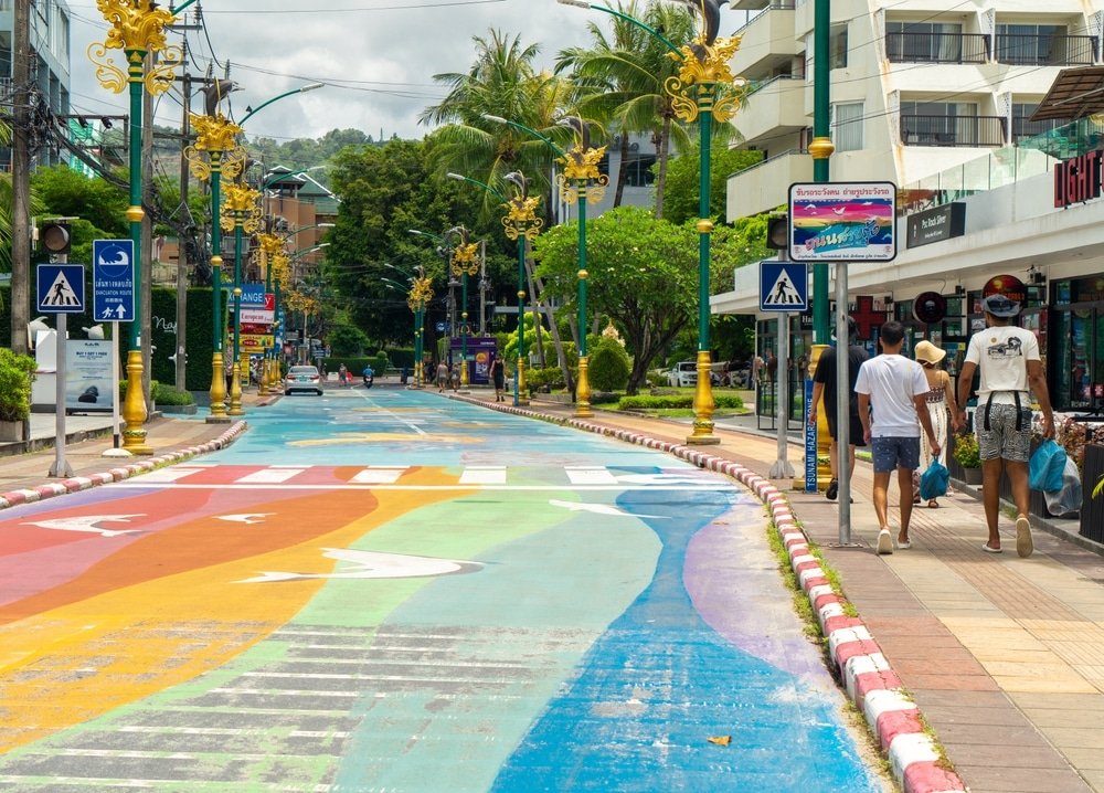 ถนนคนเดินที่ทาสีสดใสในเมืองเขตร้อนที่มีการออกแบบสีสันสดใสบนถนน เรียงรายไปด้วยต้นปาล์ม อาคารทันสมัย และป้ายจราจร ผู้คนเดินบน ที่เที่ยวภูเก็ต ทางเท้าสำรวจสถานที่ท่องเที่ยวใกล้เคียง เช่น วัดภูเก็ต