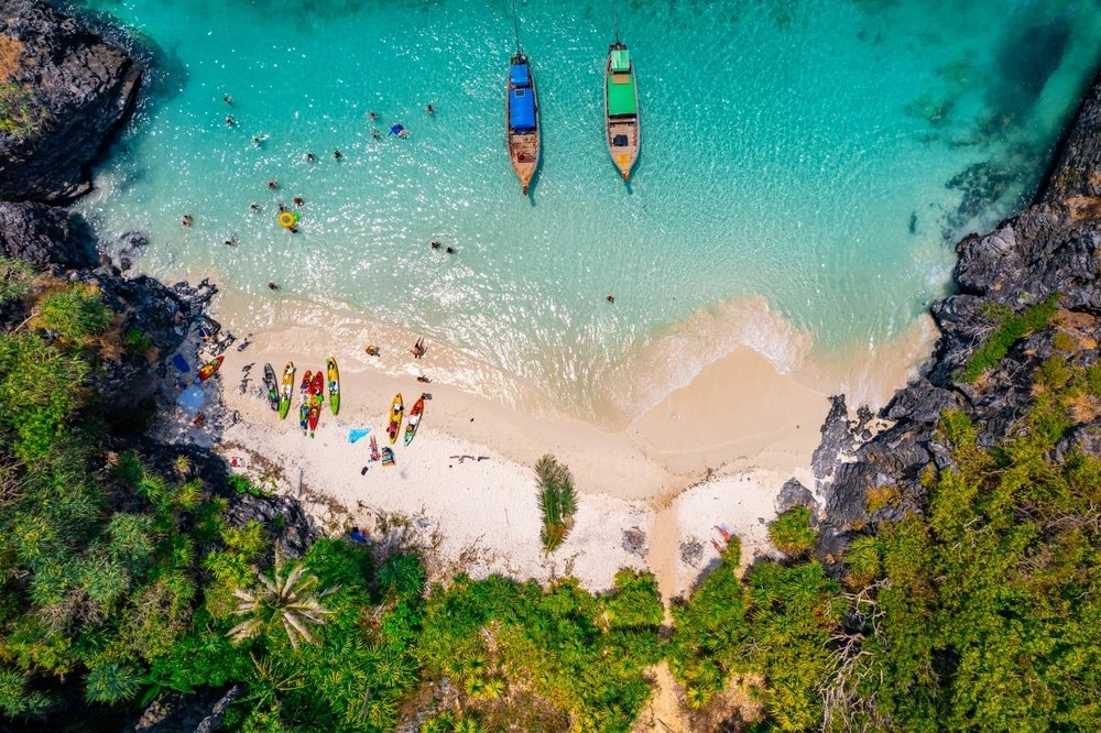 มุมมองทางอากาศของชายหาดเขตร้อนที่มีน้ำทะเลสีฟ้าครามใส ภูเก็ตที่เที่ยวภูเก็ตที่เที่ยว เรือหลายลำจอดทอดสมออยู่นอกชายฝั่ง ผู้คนกำลังว่ายน้ำและอาบแดด และเรือคายัคเรียงรายอยู่บนชายฝั่งทรายที่ล้อมรอบด้วยใบไม้สีเขียว บริเวณใกล้เคียงมีวัดภูเก็ตอันเงียบสงบเพิ่มสัมผัสทางวัฒนธรรมให้กับฉากที่งดงาม