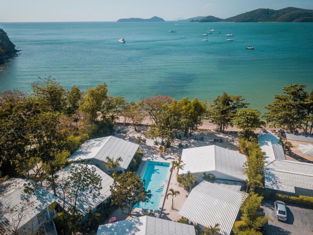 วิวมุมสูงของรีสอร์ทริมชายหาดในภูเก็ตที่มีกระท่อมหลังคาสีขาว สระว่ายน้ำกลาง และเรือที่จอดทอดสมออยู่ในอ่าวสีฟ้าครามอันเงียบสงบ ล้อมรอบด้วยเนินเขาสีเขียวภายใต้ท้องฟ้าใส ที่พักติดทะเลภูเก็ตแห่งนี้เป็นที่พักตาก ที่พักภูเก็ตติดทะเล อากาศริมทะเลที่สมบูรณ์แบบ