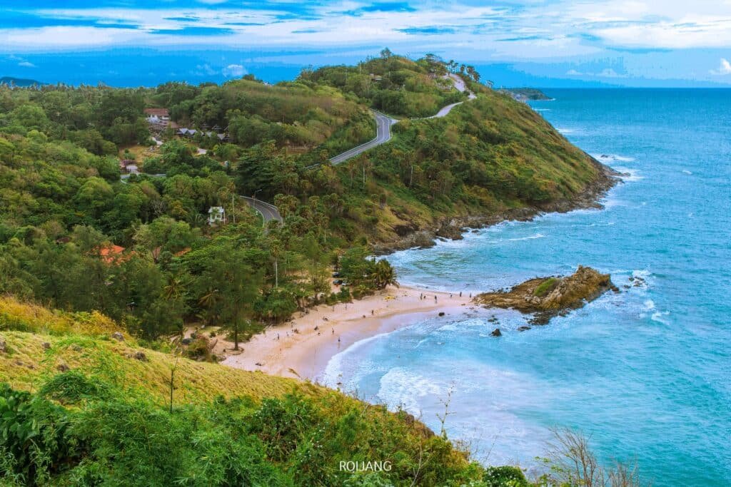 ภาพชายฝั่งทะเลแสดงให้เห็นชายหาดเล็กๆ ที่ซ่อนตัวอยู่ในอ่าว หาดยะนุ้ย ล้อมรอบด้วยเนินเขาสีเขียวและมีถนนคดเคี้ยว ผู้คนจำนวนน้อยกระจายตัวอยู่ตามชายหาดทราย ขณะที่คลื่นทะเลซัดเข้ามากระทบชายหาดอย่างแผ่วเบาใกล้แหลมพรหมเทพ