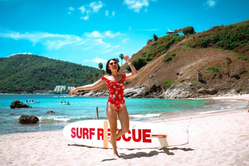 ผู้หญิงในชุดว่ายน้ำสีแดงยืนอยู่ใกล้กระดาน "Surf Rescue" บนหาดป่าตอง ซึ่งเป็นสถานที่ท่องเที่ยวยอดนิยม ที่เที่ยวป่าตอง โดยมีน้ำทะเลสีฟ้าใสและเนินเขาสีเขียวเป็นฉากหลัง