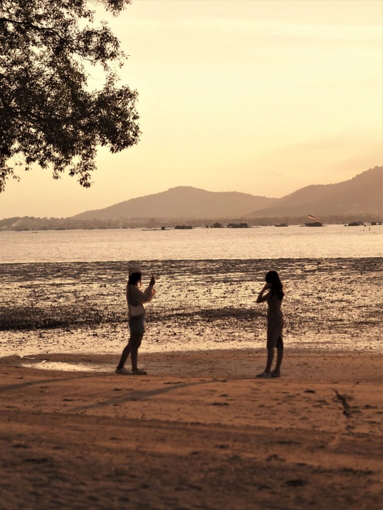 คนสองคนยืนอยู่บนชายหาดยาม  วัดฉลองภูเก็ต พระอาทิตย์ตก คนหนึ่งกำลังถ่ายรูป ในขณะที่อีกคนกำลังโพสท่า เงาของเนินเขาที่อยู่ไกลออกไปและวัดฉลองภูเก็ตปรากฏให้เห็นเหนือน้ำที่สงบนิ่งเป็นฉากหลัง