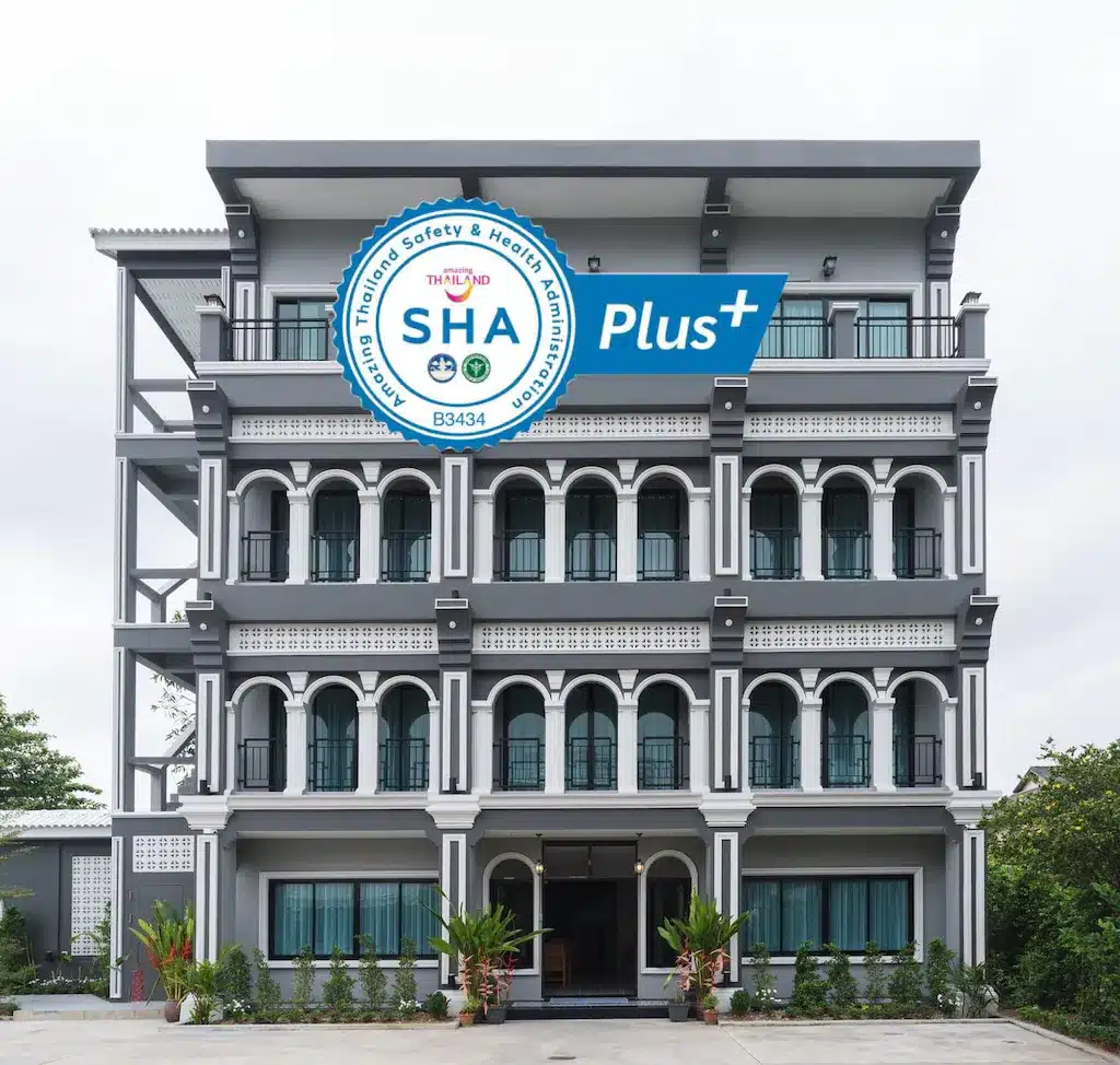 อาคารสีเทาหลายชั้นพร้อมหน้าต่างโค้ง ที่พักเมืองเก่าภูเก็ต และป้ายรับรอง "SHA Plus+" ที่ด้านหน้าอาคาร การออกแบบที่ทันสมัยของย่านนี้ในย่านภูเก็ตเสริมด้วยแมกไม้เขียวขจีโดยรอบ