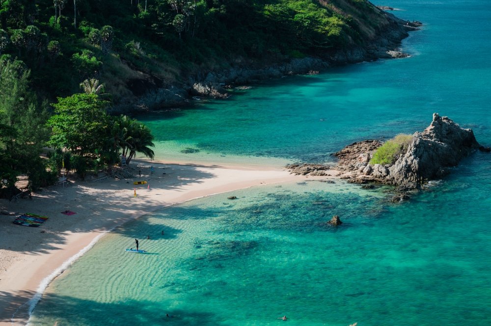 มุมมองทางอากาศของชายหาดที่เงียบสงบ มีน้ำทะเลสีฟ้าใส หาดยะนุ้ย เขียวขจี และโขดหินที่โผล่ขึ้นมาใกล้แหลมพรหมเทพ คนๆ หนึ่งกำลังยืนบนแพดเดิลบอร์ดใกล้ชายฝั่ง