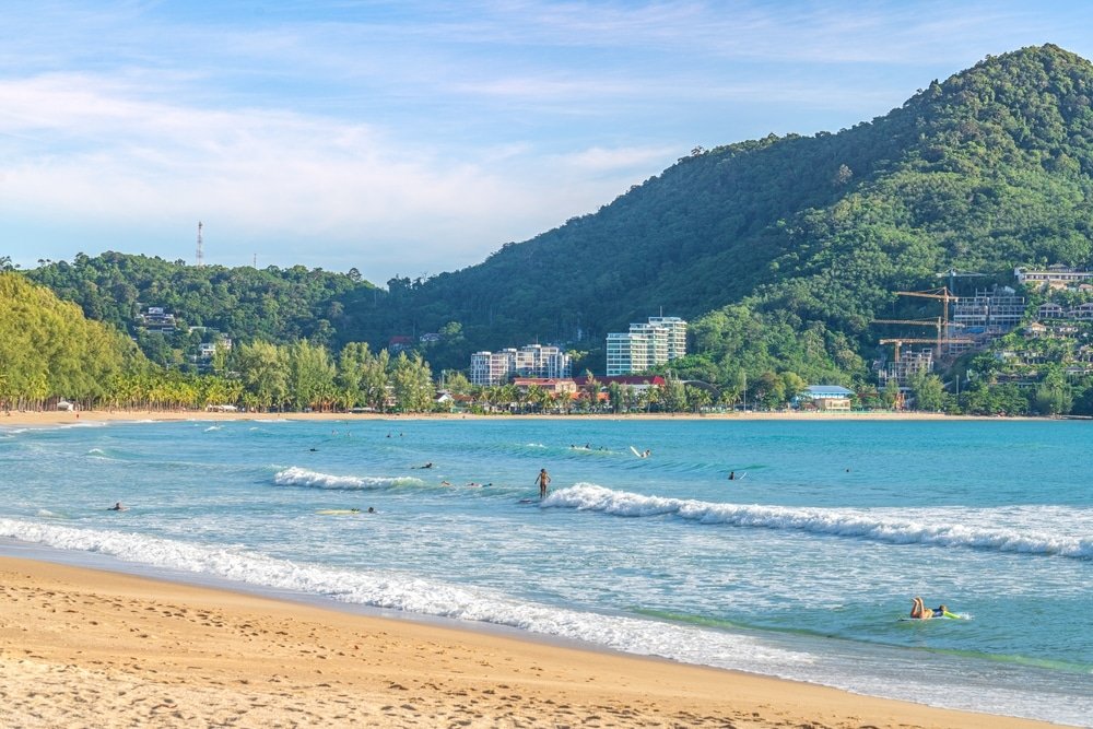 ชายหาดทรายที่มีผู้คนเล่นน้ำและเล่นเซิร์ฟ รายล้อมด้วยน้ำทะเลสีฟ้าใส ที่เที่ยวป่าตอง และเนินเขาสีเขียวขจีที่มีอาคารบ้านเรือนอยู่รายล้อม นับเป็นภาพที่สมบูรณ์แบบของ ที่เที่ยวป่าตอง