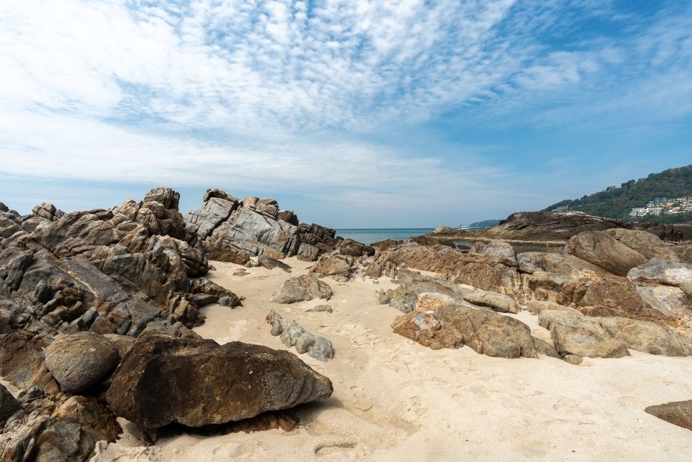 ชายหาดหินที่มีหินและทรายขนาดใหญ่ภายใต้ท้องฟ้าที่มีเมฆบางส่วน ที่เที่ยวป่าตอง เป็นสถานที่พักผ่อนอันเงียบสงบ เนินเขาและพืชพรรณกระจัดกระจายเป็นพื้นหลัง ชวนให้นึกถึงความงามตามธรรมชาติของที่เที่ยวที่