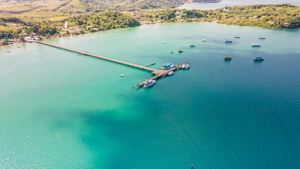 มุมมองทางอากาศของท่าเรือยาวทอดยาวสู่ทะเลสีฟ้าครามที่ชายหาดอ่าวยน มีเรือหลายลำจอดทอดสมออยู่ใกล้ๆ และมีเมืองชายฝั่งที่ล้อมรอบด้วยแมกไม้เขียวขจีเป็นฉากหลัง