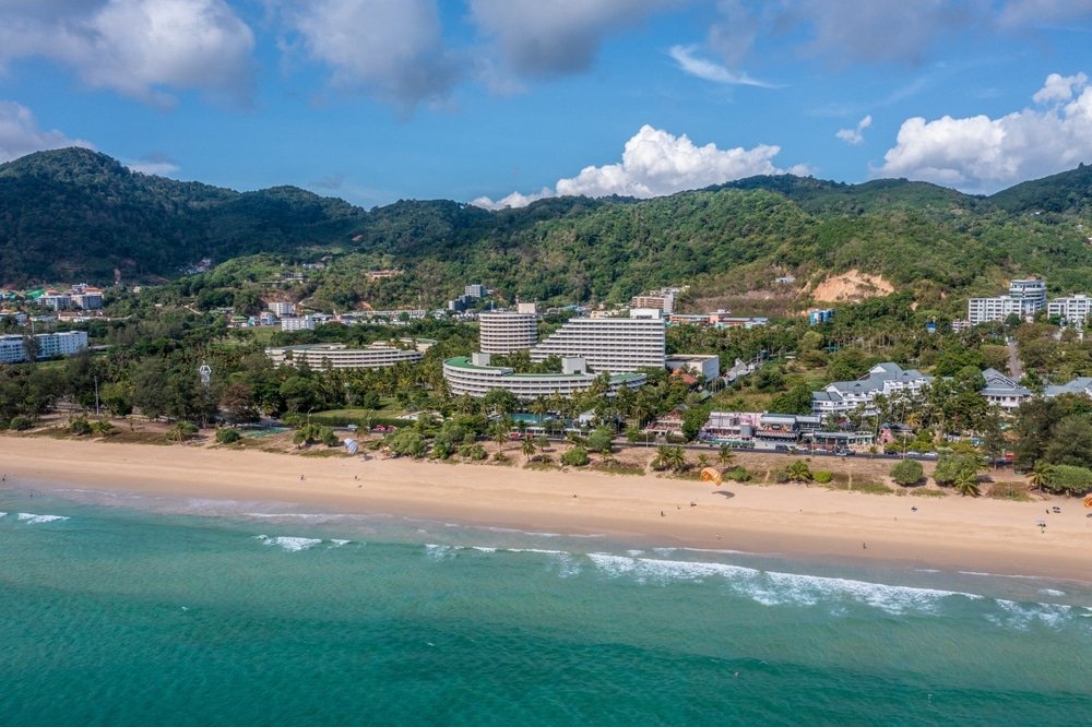 มุมมองทางอากาศของรีสอร์ทริมชายหาด ที่เที่ยวป่าตอง จัดแสดงอาคารหลายหลังที่ล้อมรอบด้วยเนินเขาสีเขียวชอุ่ม หาดทราย และน้ำทะเลสีฟ้าครามภายใต้ท้องฟ้าที่มีเมฆบางส่วน