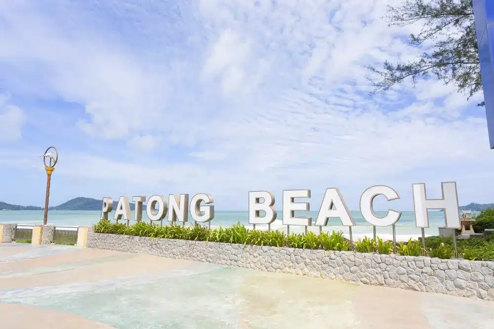 ทิวทัศน์ของหาดป่าตองที่มีตัวอักษรสีขาวขนาดใหญ่เขียนว่า “PATONG BEACH” วัดฉลองภูเก็ต โดยมีฉากหลังเป็นทะเล ท้องฟ้า และต้นไม้เขียวขจี พื้นเป็นทางลาดยาง มองเห็นเสาไฟถนนอยู่ด้านซ้าย และมีวัดฉลองภูเก็ตอยู่ด้านหลังขอบฟ้า