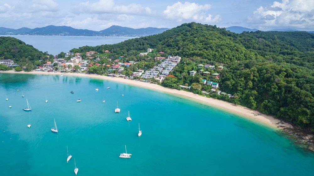 มุมมองทางอากาศของชายหาดหาดอ่าวยน ชายหาดอันเงียบสงบที่มีน้ำทะเลสีฟ้าคราม เรือใบหลายลำทอดสมอ และมีภูมิประเทศเป็นเนินเขาสีเขียวเป็นฉากหลัง