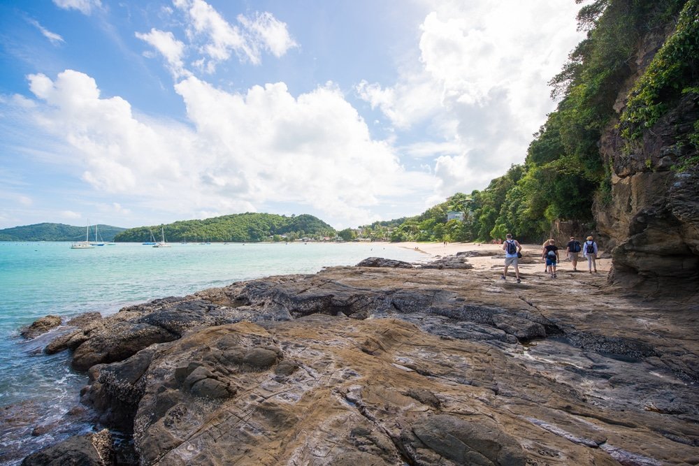 กลุ่มคนเดินไปตามแนวชายฝั่งหินของหาดอ่าวยน พร้อมทิวทัศน์ของเรือที่ทอดสมออยู่ในน้ำทะเลสีฟ้าครามและเนินเขาสีเขียวใต้ท้องฟ้าที่มีเมฆบางส่วน