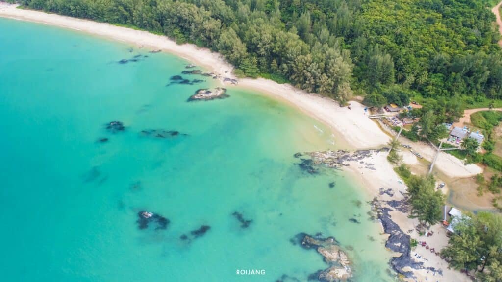 ภาพมุมสูงของชายหาดเขตร้อนในจังหวัดพังงา (เที่ยวพังงา) ที่เที่ยวพังงา ที่มีน้ำทะเลสีฟ้าใส ทรายสีขาว หินรูปร่างแปลกตา และต้นไม้เขียวขจีเป็นฉากหลัง สามารถมองเห็นอาคารเล็กๆ ใกล้ชายฝั่ง