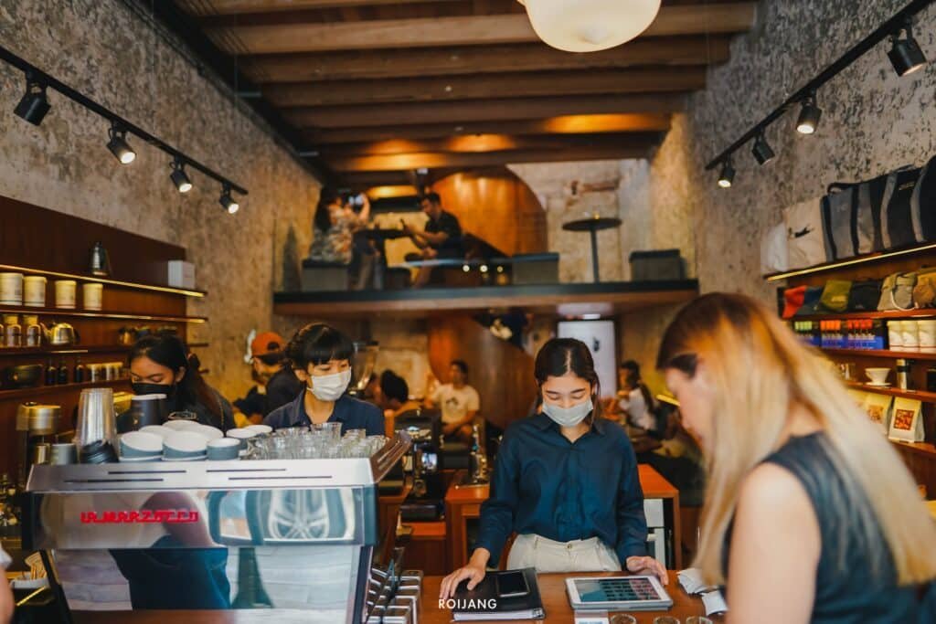 ภายในร้านกาแฟที่พลุกพล่าน โดยมีบาริสต้าทำงานอยู่หลังเคาน์เตอร์ และ  วัดพระใหญ่ภูเก็ต ลูกค้ากำลังสนทนากัน ภายใต้การจ้องมองอันเงียบสงบของรูปถ่ายในกรอบของพระพุทธมิ่งมงคลเอกนาคคีรี พื้นที่มีการออกแบบที่เรียบง่ายพร้อมชั้นวางไม้และบริเวณที่นั่งใต้หลังคา