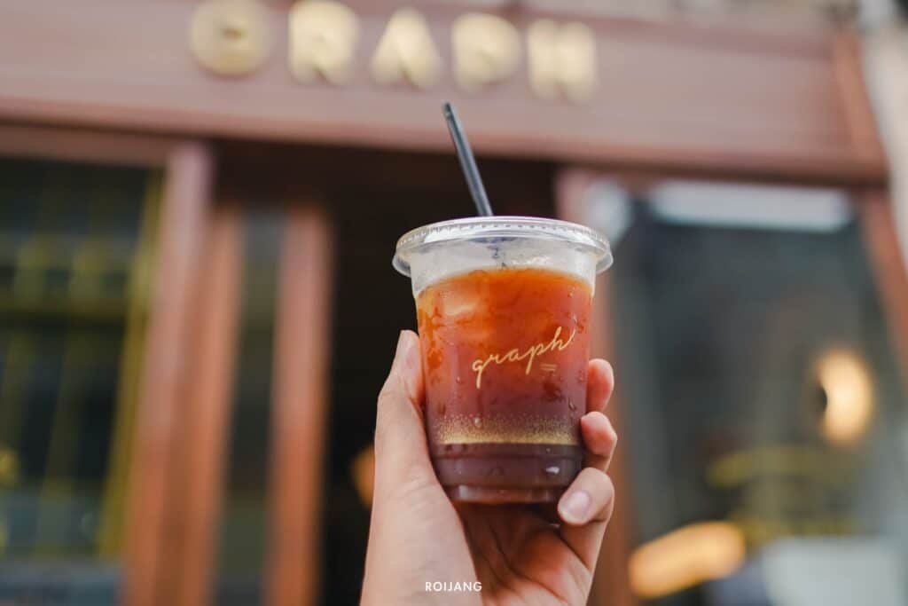 มือจับแก้วกาแฟเย็นพร้อมหลอดพลาสติกอยู่หน้าร้านกาแฟที่มีป้าย "กราฟ"  วัดพระใหญ่ภูเก็ต อยู่เหนือทางเข้า ไม่ไกลจากวัดพระใหญ่ภูเก็ต