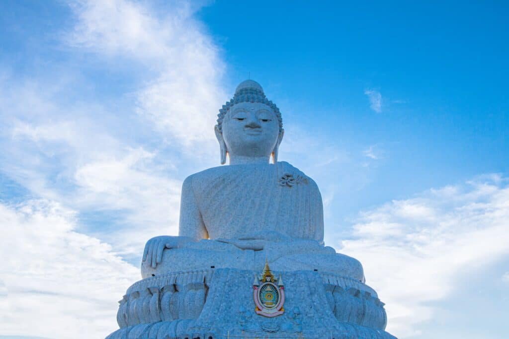 พระพุทธรูปสีขาวขนาดใหญ่ที่รู้จักกันในชื่อพระพุทธมิ่งมงคลเอกนาคคีรี ตั้งตระหง่านโดยมีท้องฟ้าสีครามและเมฆเป็นฉากหลัง ณ วัดพระใหญ่ภูเก็ต รูปปั้นมีการแกะสลักอย่างประณีตและมีสัญลักษณ์อยู่ที่ฐาน