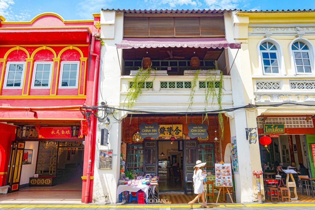 ภาพถนนของอาคารแบบดั้งเดิมสีสันสดใสพร้อมร้านค้าและร้านอาหาร โดยมีด้านหน้าอาคารเป็นสีแดง เหลือง และครีมที่สดใส คนๆ หนึ่งสวมชุดสีขาวและสวมหมวกเดินผ่านอาคารกลางเมือง ซึ่งชวนให้นึกถึงอาคารที่อยู่ใกล้ๆ  วัดพระใหญ่ภูเก็ต