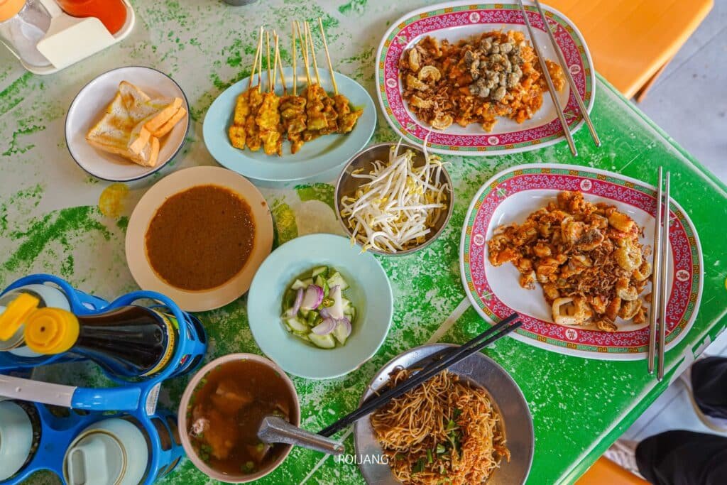 อาหารข้างทางของมาเลเซียหลากหลายรายการ เช่น สะเต๊ะ ก๋วยเตี๋ยว ผัดหมี่ และก๊วยเตี๋ยวทอด ถูกจัดวางอย่างสวยงามบนโต๊ะสีเขียวที่ร้านอาหารพื้นเมืองภูเก็ต จานขนมปังขาวและที่วางเครื่องดื่มสีฟ้าก็มองเห็นได้ ช่วยเสริมบรรยากาศ ร้านอาหารภูเก็ต ที่มีชีวิตชีวาในภูเก็ต