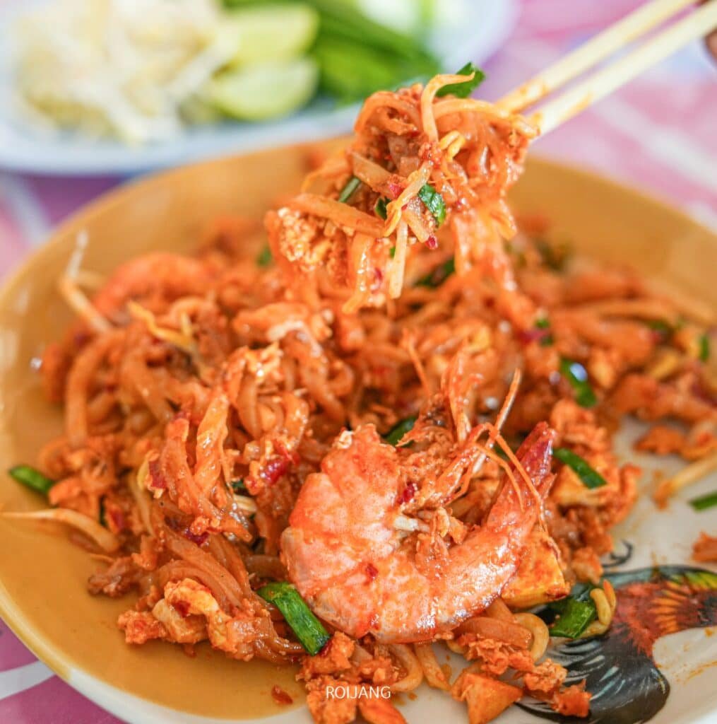 ผัดไทยใส่กุ้ง โรยหน้าด้วยผัก ตะเกียบกำลังถือส่วนหนึ่งไว้เพื่อแสดงรสชาติที่แท้จริง ร้านอาหารภูเก็ต ที่คุณสามารถพบได้ที่ร้านอาหารพื้นเมืองภูเก็ต