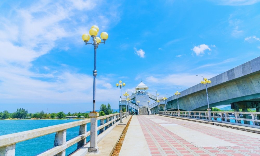 ทางเดินปูด้วยเสาไฟนำไปสู่โครงสร้างเหนือน้ำ ภายใต้ท้องฟ้าสีฟ้าที่มีเมฆ สะพานสารสิน กระจัดกระจาย ทางด้านขวามีสะพานคอนกรีตรักสารสินขนานไปกับทางเดิน