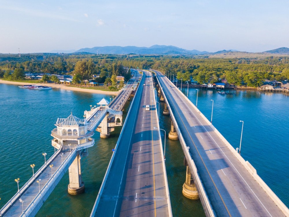 มุมมองทางอากาศของสะพาน สะพานสารสิน ที่ทอดข้ามผืนน้ำไปสู่ภูมิประเทศที่เป็นภูเขา โดยมียานพาหนะวิ่งผ่านไปตามสะพานและพื้นที่สีเขียวโดยรอบ