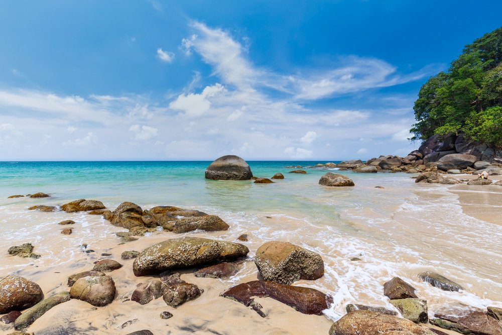 ชายหาดเขตร้อนในพังงาที่เที่ยวมีหินขนาดใหญ่กระจัดกระจายอยู่บนผืนทรายและในน้ำตื้น มหาสมุทรเป็นส่วนผสมของสีฟ้าเทอร์ควอยซ์และสีเขียว และท้องฟ้ามีเมฆบางส่วน มองเห็นต้นไม้ทางด้านขวา ที่เที่ยวพังงา ทำให้เป็นสถานที่ท่องเที่ยวพังงาที่น่าหลงใหลแห่งนี้