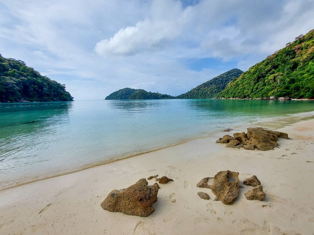 ชายหาดที่เงียบสงบมีน้ำทะเลใส ที่เที่ยวพังงา สะอาดรายล้อมด้วยเนินเขาสีเขียวขจีภายใต้ท้องฟ้าที่มีเมฆบางส่วน มีหินกระจัดกระจายอยู่ตามชายหาดทราย ทำให้เป็นจุดที่สมบูรณ์แบบสำหรับการสำรวจอัญมณีที่ซ่อนเร้นแห่งหนึ่งในพังงาที่เที่ยว