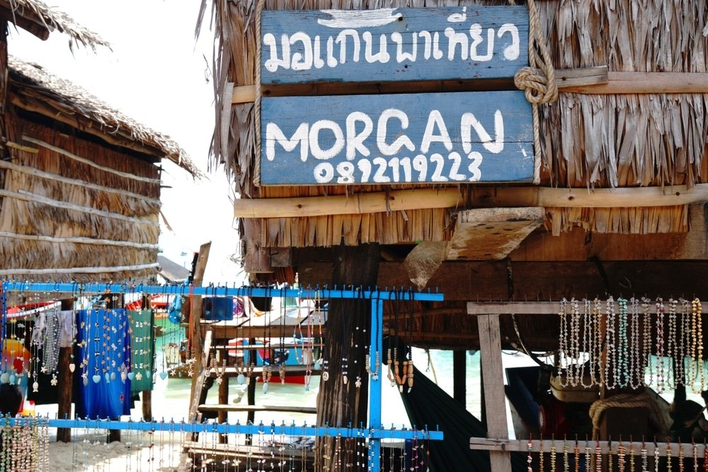 ร้านค้าไม้ริมชายหาดที่มีหลังคาฟาง มีป้ายที่เขียนทั้งภาษาไทยและภาษาอังกฤษพร้อมหมายเลขโทรศัพท์ ล้อมรอบด้วยเครื่องประดับลูกปัดห้อยระย้า เป็นจุดแวะพักที่สมบูรณ์แบบสำหรับผู้ที่มาสำรวจ พังงาที่เที่ยว