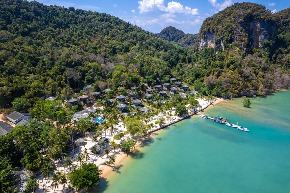 มุมมองทางอากาศของรีสอร์ทริมชายหาดในเขตร้อนที่มีบังกะโลตั้งอยู่ท่ามกลางต้นไม้เขียวขจี ที่เที่ยวพังงา รายล้อมด้วยน้ำทะเลสีฟ้าใส ชายหาดทราย และหน้าผาที่มีป่าไม้ภายใต้ท้องฟ้าที่มีเมฆบางส่วน เป็นจุดหมายปลายทางที่งดงามและเงียบสงบ ที่เที่ยวพังงา