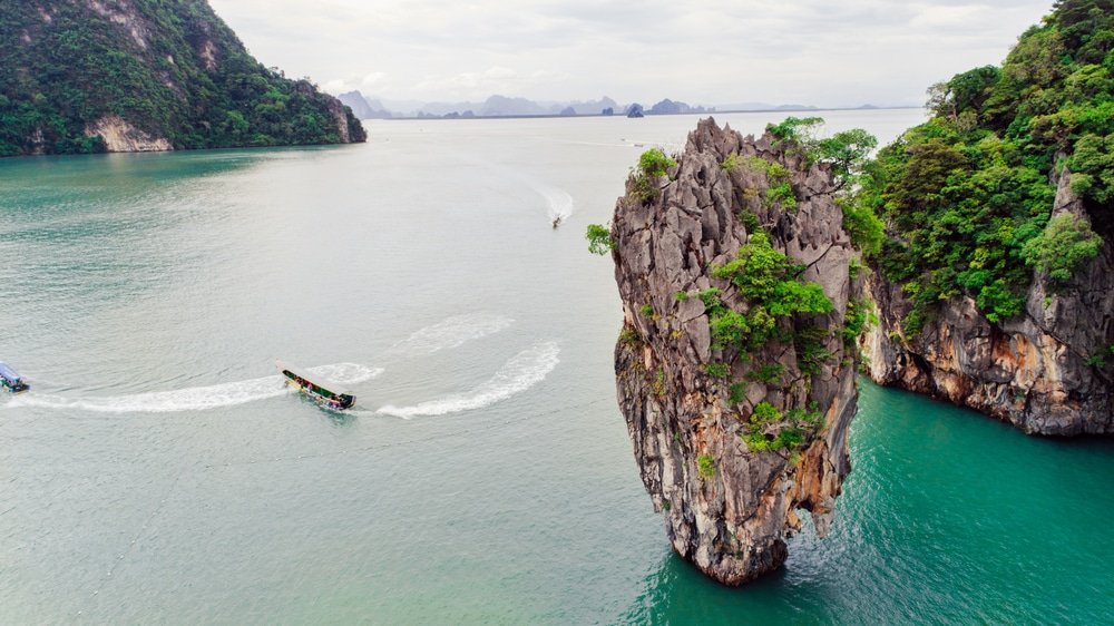 ภาพมุมสูงของหินปูนขนาดใหญ่ที่ปกคลุมไปด้วยต้นไม้สีเขียวในน้ำทะเลสีฟ้าอมเขียวที่สงบ พร้อมเรือที่มองเห็นได้ในน้ำ และเนินเขาเขียวขจีเป็นฉากหลัง แสดงให้เห็นความงามที่น่าทึ่งของ เกาะตะปู