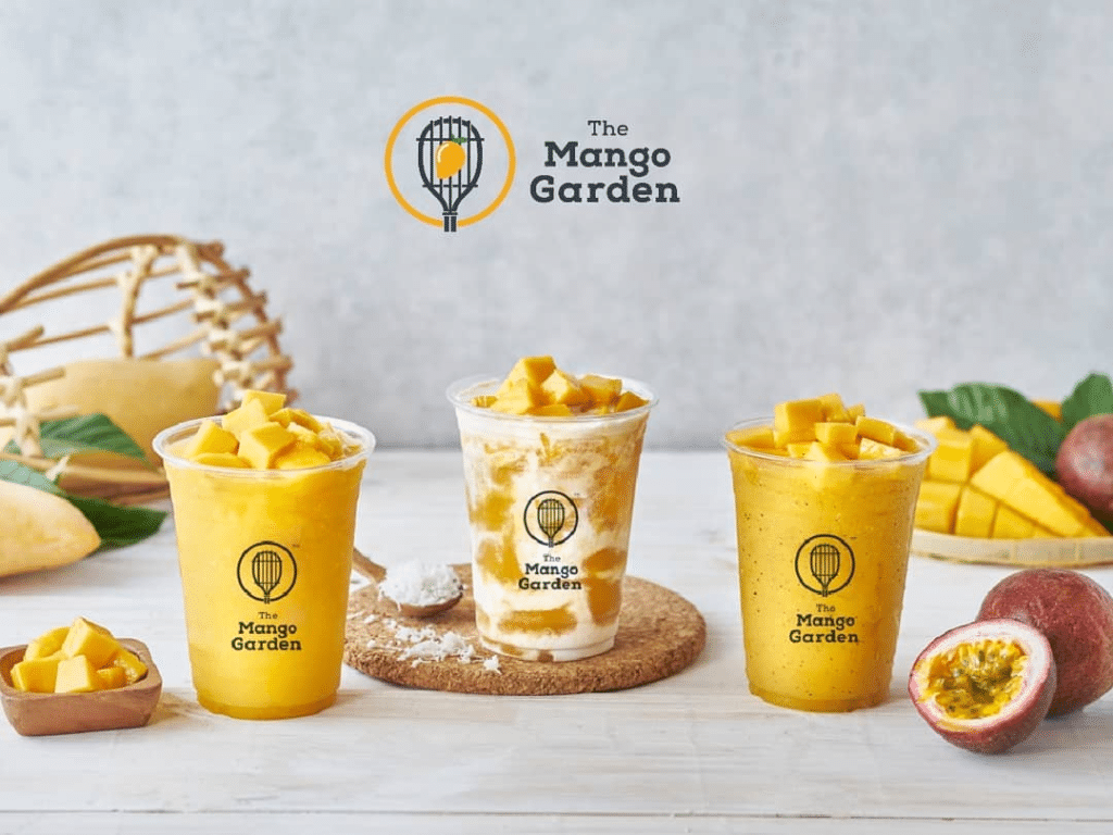 เครื่องดื่มธีมมะม่วง 3 ชนิดในแก้วใส พร้อมโลโก้ The Mango Garden ประดับด้วยชิ้นมะม่วง และล้อมรอบด้วยมะม่วงสด เสาวรส และใบไม้เมืองร้อนบนพื้นผิวสีขาว ชวนให้นึกถึงกลิ่นหอมสดชื่นของ เกาะพีพี ในทุกจิบ