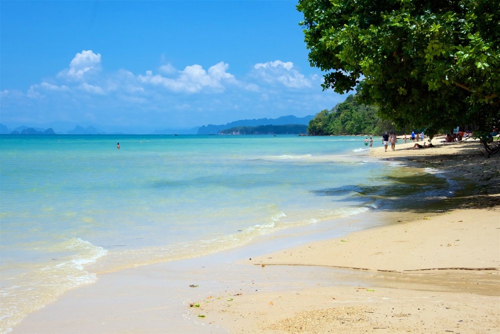 หาดทรายในจังหวัดกระบี่ที่มีคลื่นอ่อนโยนและน้ำทะเลสีฟ้าครามภายใต้ ที่เที่ยวกระบี่ ท้องฟ้าสีฟ้าใส ผู้คนต่างเดินพักผ่อนริมน้ำ โดยมีต้นไม้เขียวขจีให้ร่มเงาทางด้านขวา เหมาะสำหรับใครก็ตาม ดูเที่ยวกระบี่ หรือ ท่องเที่ยวกระบี่