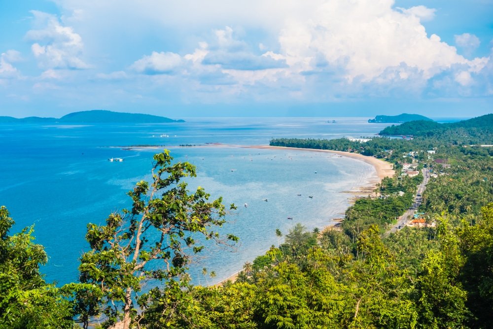 ภาพมุมสูงของชายฝั่งทะเลเขตร้อนที่มีหาดทราย ที่เที่ยวชุมพร ต้นไม้เขียวชอุ่ม และเรือบนมหาสมุทรสีฟ้าภายใต้ท้องฟ้าที่มีเมฆบางส่วน มองเห็นเกาะต่างๆ อยู่ไกลๆ แสดงให้เห็นว่าชุมพรที่เที่ยวเป็นจุดหมายปลายทางในอุดมคติสำหรับผู้รักธรรมชาติ