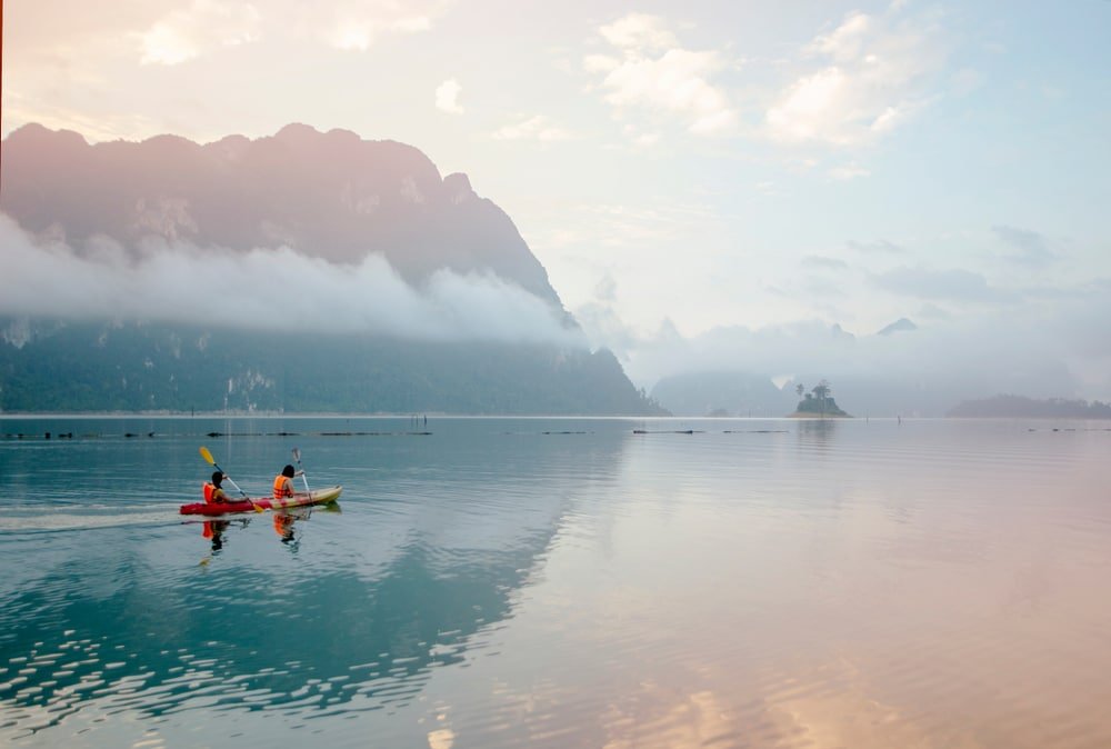 คนสองคนกำลังพายเรือคายัค เขื่อนเชี่ยวหลาน ในทะเลสาบอันเงียบสงบที่เขื่อนรัชชประภา โดยมีภูเขาหมอกเป็นฉากหลังภายใต้ท้องฟ้าที่มีเมฆมาก