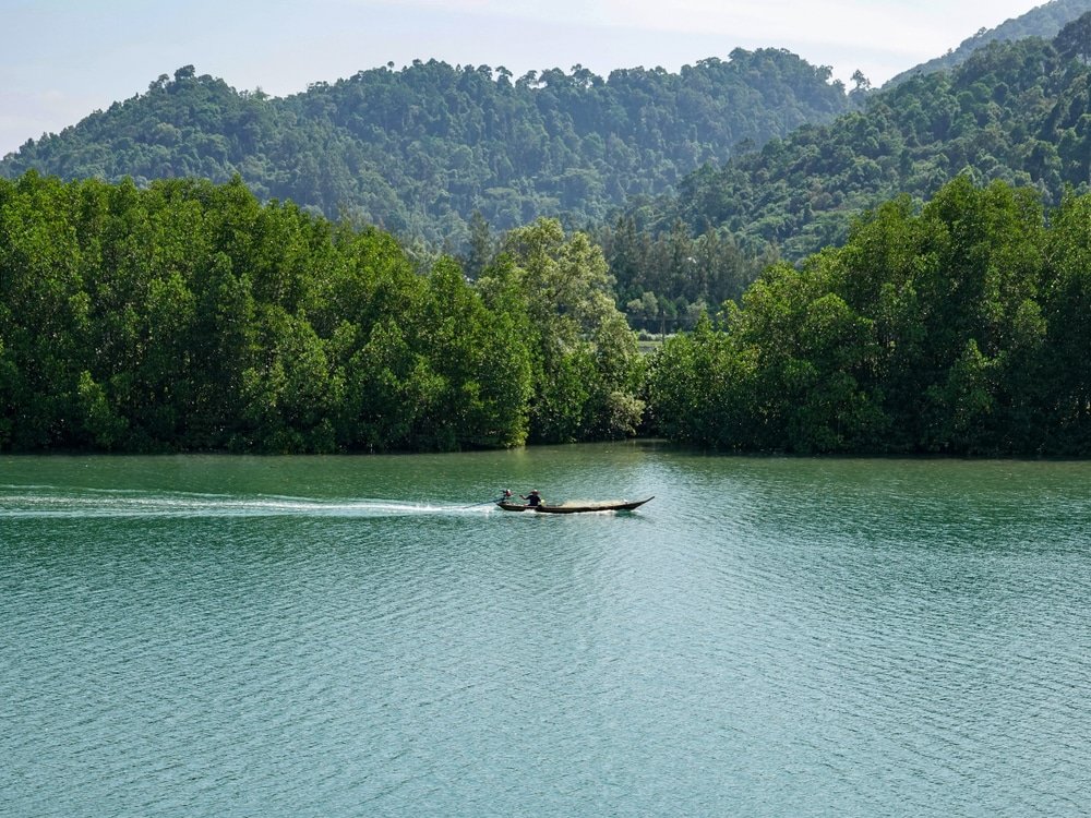 เรือเล็กพร้อมคนสองคนล่องไปใน ที่เที่ยวชุมพร แม่น้ำอันเงียบสงบในจังหวัดชุมพร รายล้อมไปด้วยต้นไม้สีเขียวชอุ่มและเนินเขาไกลๆ ใต้ท้องฟ้าใส จับภาพความงามอันเงียบสงบของ ที่เที่ยวชุมพร