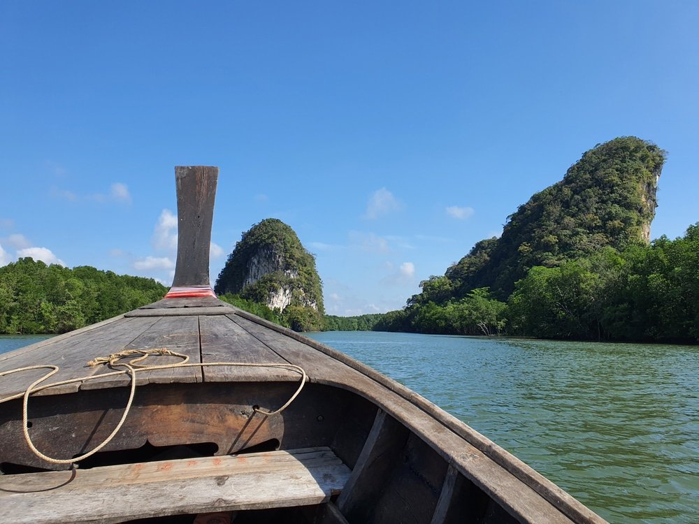 เรือไม้ลอยอยู่ในแม่น้ำอันเงียบสงบที่ล้อมรอบด้วยเนินเขาสีเขียวชอุ่มภายใต้ท้องฟ้าสีฟ้าใส ท่องเที่ยวกระบี่ ชวนให้นึกถึงทิวทัศน์อันงดงามที่พบในรีวิวเที่ยวกระบี่