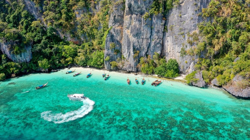 มุมมองทางอากาศของชายฝั่งเกาะพีพีอันสวยงามตระการตา พร้อมด้วยน้ำทะเลสีฟ้าใส เรือที่ทอดสมออยู่ใกล้ เกาะพีพี ชายหาด และหน้าผาสูงชันที่ปกคลุมไปด้วยต้นไม้เขียวขจีเป็นฉากหลัง