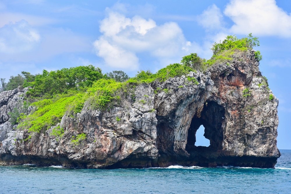 เกาะหินที่มีรูปร่างโค้งตามธรรมชาติ  ที่เที่ยวชุมพร ปกคลุมไปด้วยพืชพรรณสีเขียว ล้อมรอบด้วยน้ำทะเลสีฟ้าภายใต้ท้องฟ้าที่มีเมฆบางส่วน เป็นทัศนียภาพอันน่าทึ่งแห่งหนึ่งที่คุณจะได้พบขณะสำรวจชุมพรที่เที่ยว